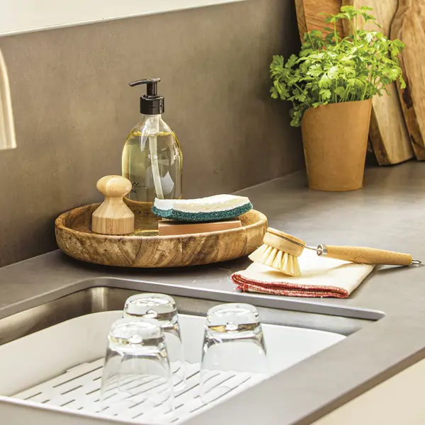 fregadero de cocina, jabón, cepillo y esponja, vasos tablas de madera, planta aromática, ventana con estor